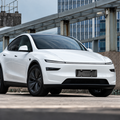 A white Tesla Model Y is shown from a front three-quarter angle on an outdoor rooftop with a glass office building in the background.