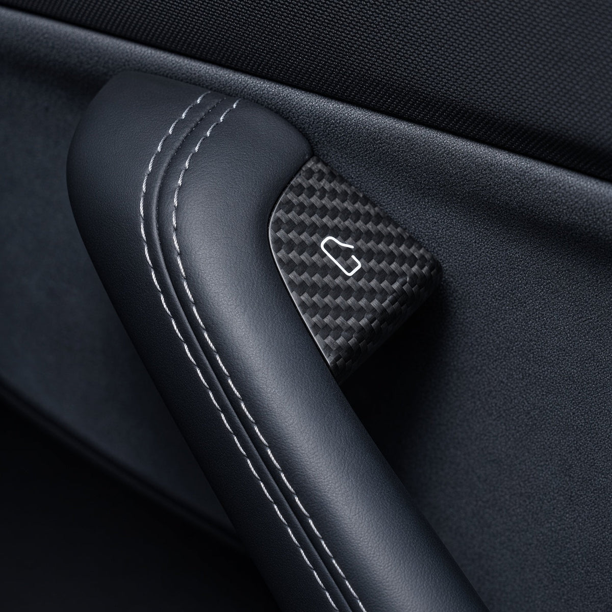 Close-up of a carbon-weave door-release cover with a white door symbol beside a stitched black grab handle.